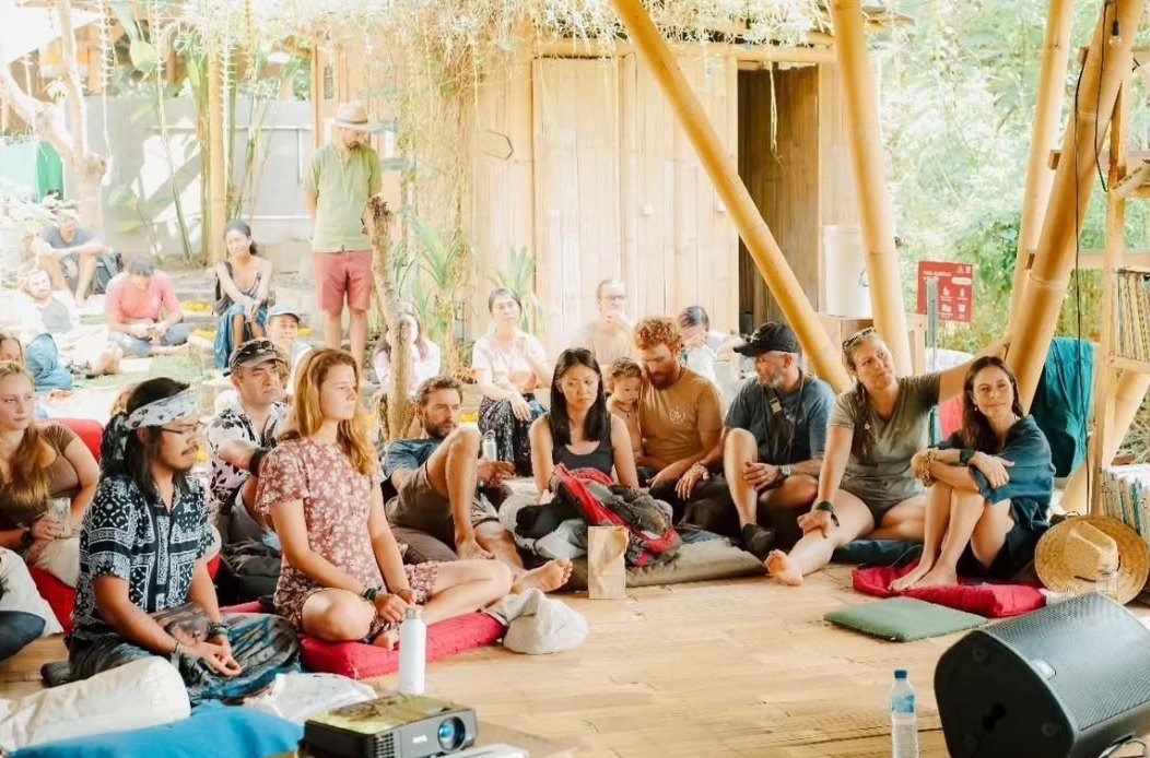 People seated on cushions in an open bamboo pavilion, listening to a talk